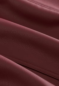 Burgundy színű műbőr anyag, amelynek texturált felülete van, finom redőkkel és varrásokkal az élek mentén. Puha és hajlékony.