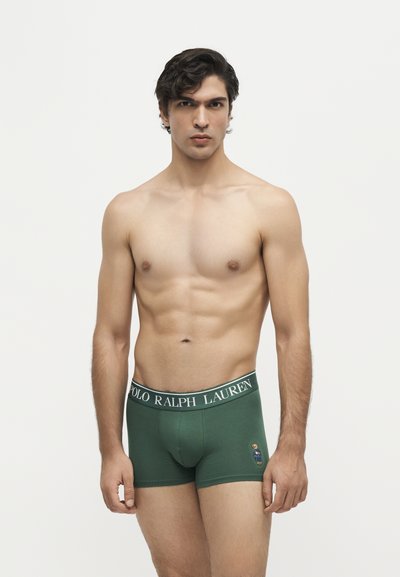 Мъжки модел, облечен в зелени боксерки Polo Ralph Lauren, стои пред едноцветен светъл фон, гледайки напред с неутрално изражение.