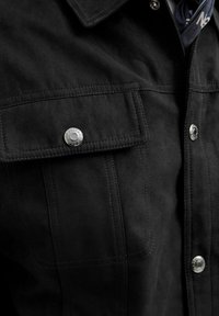 Czarna zamszowa kurtka z dwoma kieszeniami na piersi, srebrnymi zatrzaskami i widocznym przeszyciem. Miękka w dotyku, z kołnierzem.