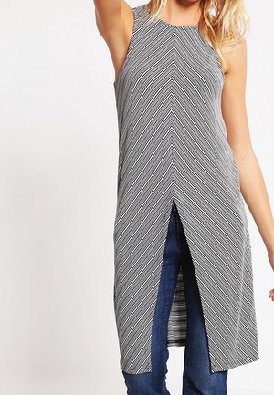 Kobieta w szarej bezrękawnikowej tunice z ukośnymi paskami i przednim rozcięciem, w połączeniu z niebieskimi dżinsami, pozująca na białym tle.