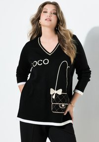 Suéter negro de punto con escote en V, acentos blancos, bordado "COCO" y un diseño de bolso acolchado beige con un lazo. Mangas largas.