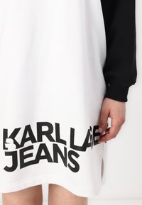 Vestido branco com mangas pretas e um grande logo preto "KARL LAGERFELD JEANS" na bainha. Material em mistura de algodão com fendas laterais.