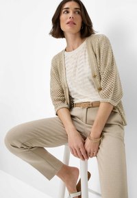 Beige Strickjacke mit lockerer Passform und offenem Gewebe. Kombiniert mit einem gestreiften Metallshirt und beigefarbenen strukturierten Hosen, accessoriert mit Armbändern.