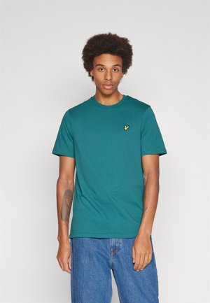 T-shirt teal in cotone con scollatura rotonda e maniche corte. Presenta un piccolo logo giallo sul lato sinistro del petto. Indossato con jeans blu.