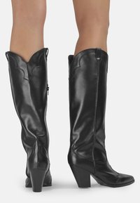 Botas altas de cuero negro hasta la rodilla con puntera afilada, tacón angular, bordes superiores ondulados y cremalleras laterales para facilitar el uso.