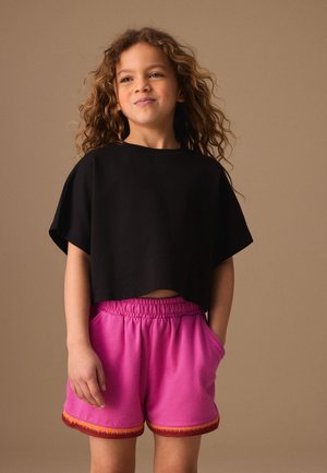 Kind met krullend haar draagt een zwart cropped t-shirt en felroze shorts met oranje bies, staand tegen een effen bruine achtergrond.