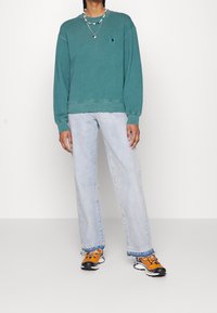 Sudadera de algodón azul verdoso con puños y dobladillo acanalados, combinada con vaqueros de mezclilla azul claro. El calzado presenta detalles en naranja y texturas mixtas.