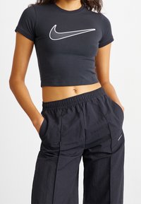 Kvinna klädd i en svart kort t-shirt med en vit Nike-logotyp och lösa svarta byxor med elastisk midja, händerna i fickorna, mot vit bakgrund.