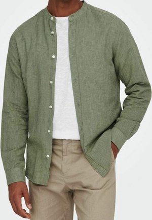 Homme portant une chemise vert clair boutonnée sur un t-shirt blanc avec un pantalon beige, main dans la poche devant un fond uni.