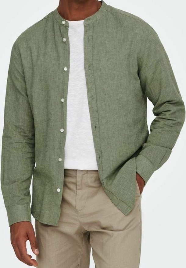 Homme portant une chemise vert clair boutonnée sur un t-shirt blanc avec un pantalon beige, main dans la poche devant un fond uni.