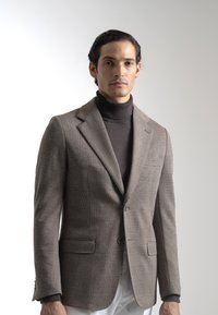 Blazer marrón a cuadros con solapas en pico, dos bolsillos frontales y cierre de un solo botón, combinado con un suéter de cuello alto oscuro.