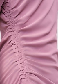 Απαλό ροζ ύφασμα με κάθετο ρυτσίνισμα που δημιουργεί πτυχωτή, συσσωρευμένη υφή κατά μήκος μιας πλευράς, αναδεικνύοντας τη λεπτομερή ραφή και το λείο υλικό.