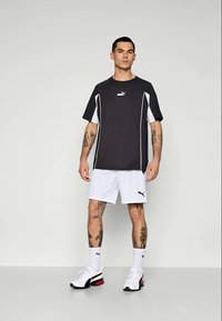 Muž stojící v černobílé sportovní výbavě Puma, v bílých ponožkách Puma a bílých teniskách Puma, s několika tetováními na pažích a nohách.