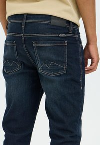 Tamnoplave traperice s uskim krojem, s dva stražnja džepa s prošivom u obliku planine i kožnim zakrpom na pojasu.