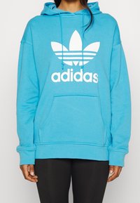 Blå huva tröja i mjukt tyg med en stor vit Adidas-logotyp och randdesign, samt en känguruficka framtill.