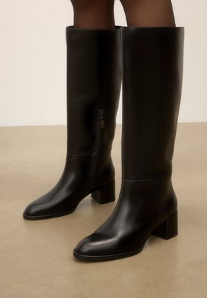 Bottes en cuir noires montant jusqu'aux genoux avec talons blocs et fermetures éclairs latérales, portées sur jambes nues contre un sol et un mur beiges unis.