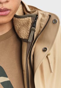 Beige Jacke mit einem weichen, beigen Fleece-Futter und einem glatten Außenmaterial. Verfügt über einen seitlichen Reißverschluss und einen Knopfverschluss als Detail.