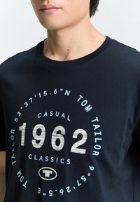 Camiseta de algodón azul marino con un gráfico redondo que presenta el texto blanco "1962", "TOM TAILOR" y coordenadas, y un suave cuello redondo.