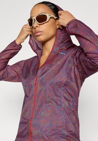 Chaqueta ligera con capucha, de color rojo y azul con un patrón, que cuenta con un cierre frontal y un logo bordado en el pecho.