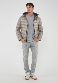 Giovane uomo che indossa una giacca a vento beige sopra una maglietta grigia, jeans azzurri e sneakers bianche con dettagli blu navy, in piedi con le mani nelle tasche.
