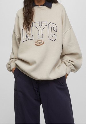 Person iført beige oversized sweatshirt med teksten "NYC" og navy brede bukser, hænderne i lommerne, foran en ensfarvet baggrund.