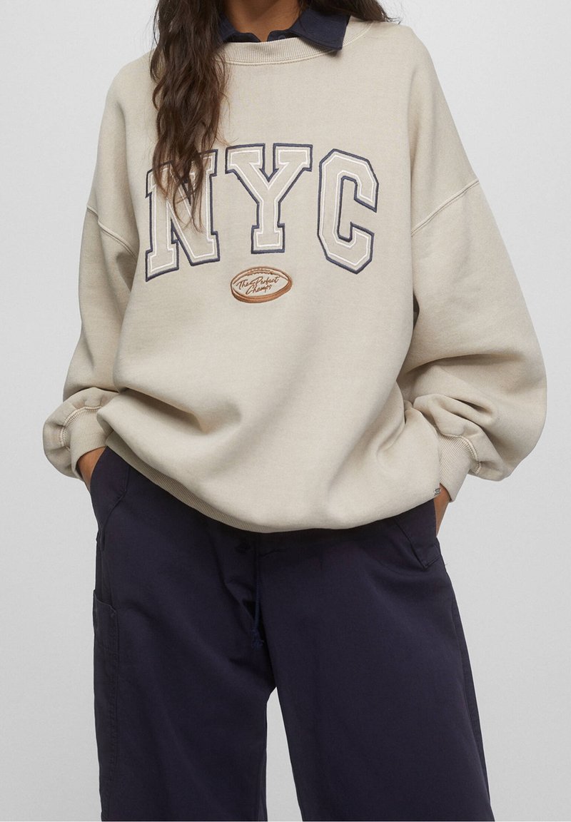 Personne portant un sweat-shirt beige oversize avec le texte "NYC" et un pantalon large bleu marine, les mains dans les poches, devant un fond uni.