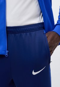 Άτομο που φοράει μπλε αθλητική φόρμα Nike με λευκό λογότυπο Swoosh, λευκό πουκάμισο και μπλε μπουφάν με το χέρι στην τσέπη.