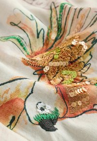 Közelről készült kép egy világos alapon látható, narancssárga, zöld, arany és fehér árnyalatú hímzett és flitteres virágmintával díszített textilről.