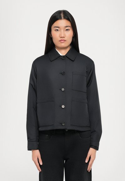 Frau mit langen, glatten Haaren, die eine geknöpfte schwarze Jacke mit drei Fronttaschen und schwarze Hosen trägt, steht vor einem weißen Hintergrund.