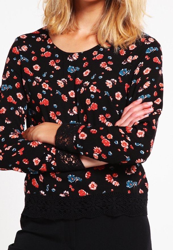Femme portant un haut noir à manches longues avec un motif floral rouge, rose et bleu et des détails en dentelle aux poignets et à l’ourlet, bras croisés.