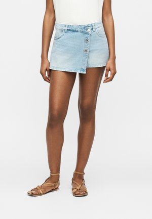 Frau trägt einen hellblauen asymmetrischen Denim-Minirock mit Knöpfen vorne, weißes Top und braune flache Sandalen mit Riemen.