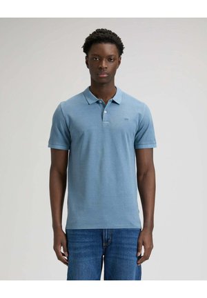 Junger Mann steht, trägt ein hellblaues Poloshirt und blaue Jeans vor einem schlichten hellen Hintergrund.