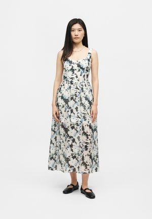 Γυναίκα με μακριά μαύρα μαλλιά που φοράει αμάνικο φόρεμα μέσης μήκους με λουλουδάτο μοτίβο και μαύρα παπούτσια Mary Jane, στέκεται μπροστά σε απλό φόντο.