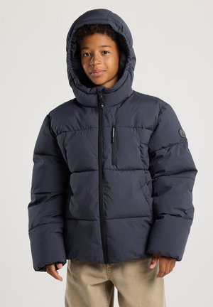 Jeune personne portant une doudoune bleu foncé avec capuche, poche poitrine zippée et pantalon beige, se tenant contre un fond uni.