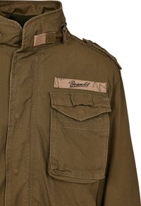 Brown military-style jacket with a high collar, front flap pockets, and Brandit label. Features textured fabric and shoulder epaulets.