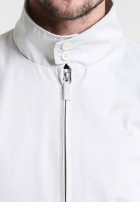 Dolna część twarzy i szyi mężczyzny noszącego białą, wysoką kurtkę z zamkiem błyskawicznym, z zapiętymi dwoma białymi guzikami przy kołnierzu.