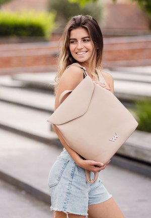 Beige leather backpack with a structured design, flap closure, and silver logo detail. Features dual handles and smooth texture.