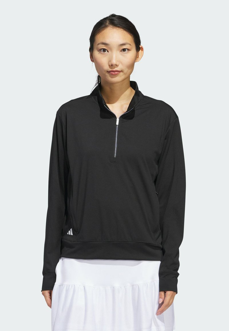 Femme debout, portant un haut noir à manches longues zippé avec une jupe blanche, sur un fond gris clair uni.