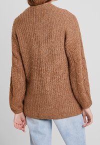 Knitted brown sweater with a ribbed texture, wide sleeves, and cable-knit accents on the arms. Soft, cozy material, length below the hips.