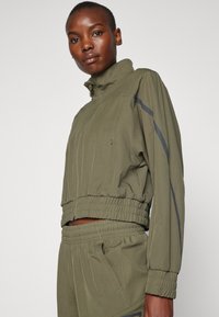 Kort olivengrønn jakke med høy krage, elastiske mansjetter og cropped passform. Har mørkegrå striper langs ermene. Glatt tekstur.