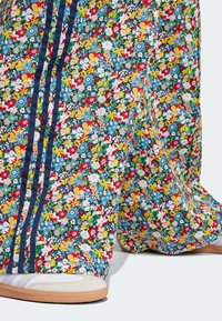 Pantaloni tuta floreali in tessuto multicolore con piccoli fiori, strisce blu scuro ai lati e una vestibilità morbida sopra scarpe bianche.