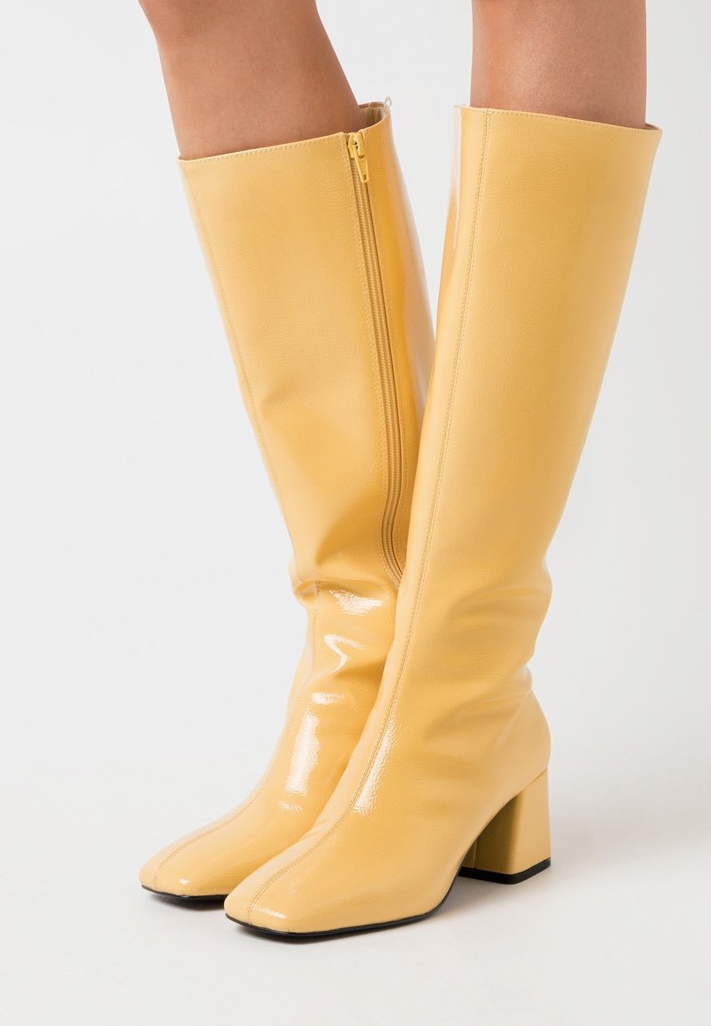 Bottes jaunes jusqu'aux genoux en matériau brillant, avec un bout carré et un talon bloc. Équipées d'une fermeture éclair sur le côté pour un enfilage facile.