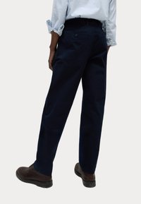 Pantaloni di cotone blu navy a gamba dritta, con tasche posteriori e chiusura a bottone. Indossati con scarpe marroni, mostrando le maniche della camicia arrotolate.