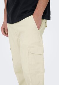Pantalon cargo beige avec poches latérales et détails cousus, associé à une chemise noire, mettant en valeur une coupe décontractée et une texture douce.