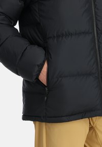 Schwarze Daunenjacke mit einem gesteppten Design, ausgestattet mit einer seitlichen Reißverschlusstasche und einer glatten Textur. Die Bündchen sind ebenfalls gepolstert für zusätzliche Wärme.