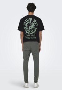 T-shirt in cotone nero con grande testo verde sul retro: "SII SOLO TE STESSO" insieme a un simbolo yin-yang. Abbinato a pantaloni grigi e sneakers bianche.