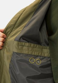 Wasserabweisende, schmutzresistente Jacke mit grünem Außenmaterial und grauem Innenfutter. Verfügt über eine glatte Textur und ein sichtbares Pflegehinweissymbol.