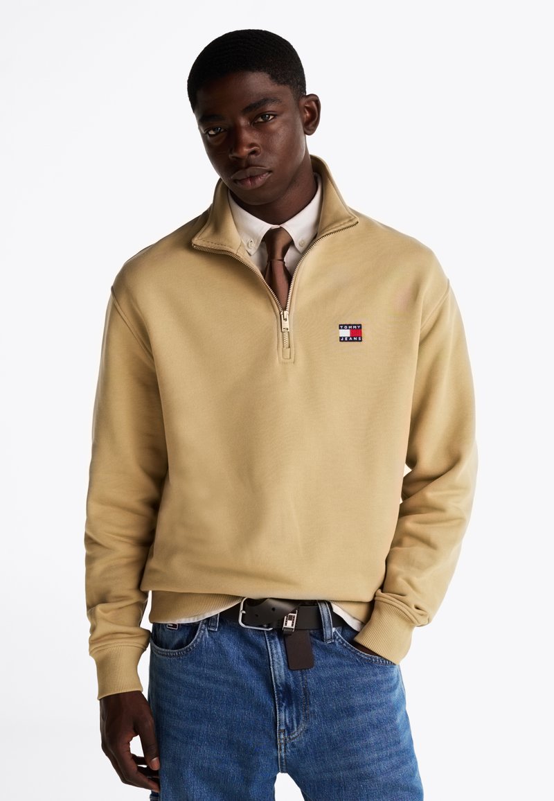 Felpa beige con zip realizzata in misto cotone, caratterizzata da un piccolo logo ricamato, polsini a costine e una vestibilità comoda, abbinata a jeans blu.