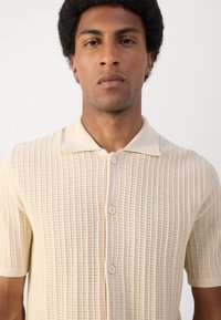 Beige knitted short-sleeve shirt with a button-up front, featuring a textured pattern and a classic collar. Smooth fabric with even stitch design.