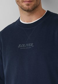 Uomo con un maglione blu navy con il logo "S.OLIVER ELEVATED SINCE 1969" sul petto, che mostra parzialmente il viso e il collo.